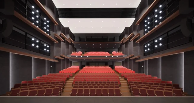 This is an image of the inside of a theater, looking out from the stage toward the audience seating. The seating area is divided into two levels: the main floor and a second-floor balcony. On the main floor, rows of red seats are arranged in a stepped, tiered layout rising from front to back. Toward the upper rear of the main floor, there is a second-floor section that projects outward like a balcony, with seats also set in a stepped arrangement from front to back. Most of the ceiling is white, with part of it forming a black horizontal band. The theater’s interior walls are gray, and on both the left and right sides there are spotlights and linear lights, along with occasional decorative elements made of wooden panels. The overall atmosphere is subdued and sophisticated.
