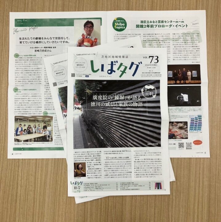 木目の机の上に、港区芝地区の地域情報誌「しばタグ」が３部少しずらして重ねて置いてあります。一番上に置いてある冊子の表紙には、「しばタグ」という大きな題字が書かれています。その下には、道路脇にある石でできた塀が広範囲に写った風景写真と記事が配置されています。一番下に置いてある冊子は見開きになっていて、みなと芸術センター開館準備室の宮崎室長の顔写真とインタビュー記事の一部のほかに、イベント風景写真などが写っています。