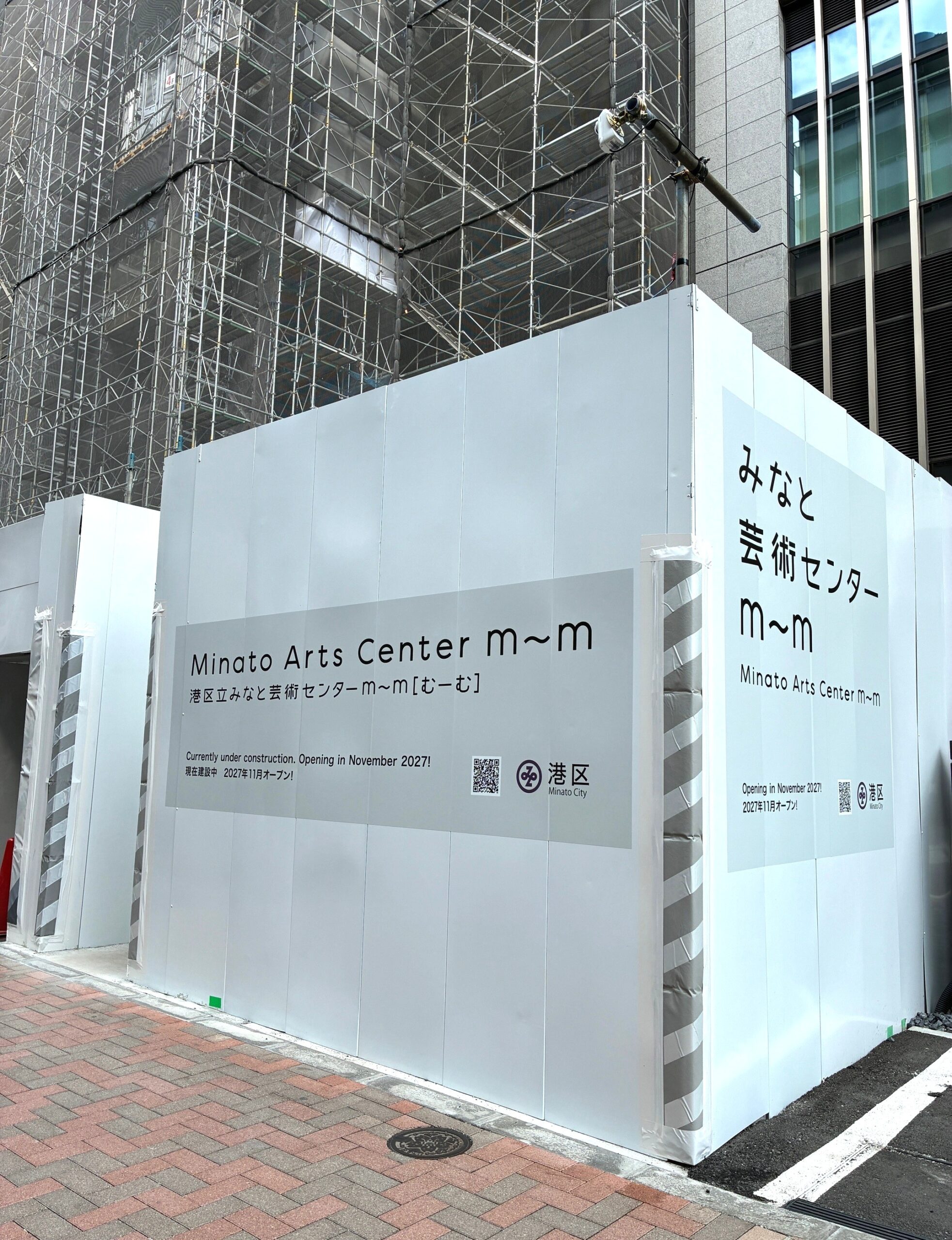 街角の歩道に面した工事現場の画像です。奥にある建築中の建物は、建物全体が足場と黒い養生ネットで覆われています。手前には白い仮囲いが立っています。仮囲いには、灰色の大きなシールが左と右にそれぞれ1枚ずつ貼ってあります。どちらのシールにも「Minato Arts Center m～m」「港区立みなと芸術センターm～m（むーむ）」、「Currently under construction.Opening in November 2027 !」「現在建設中 2027年11月オープン！」と書かれています。シールの右下には、二次元コードと港区Minato Cityと書かれたロゴマークが表示されています。