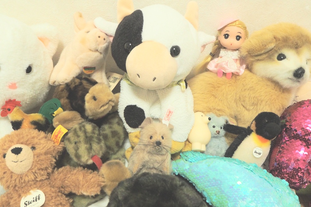 たくさんのぬいぐるみが壁際にぎゅっと集めて並べられた画像です。中央に白黒模様の牛、右に犬やペンギン、左下にクマなどが見え、前景には青緑やピンクのキラキラしたクッションのようなものもあります。右上には金髪で目が大きい小さな人形もあります。にぎやかな印象です。