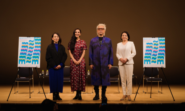 「開館２年前プロローグイベント」で行ったトークイベントのいち場面です。舞台上には、イベント出演者４人が横一列に立っています。出演者は、左から、相馬千秋、サヘル・ローズ、やない道彦、せいけ愛です。その左右には、ｍチルダｍを波模様のようにデザインしたポップな印象のポスターがイーゼルで立てられています。その後ろには、黒いパイプ椅子が数脚のほかに、ペットボトルが数個置かれたサイドテーブルがあります。舞台上は暗めの照明で照らされています。