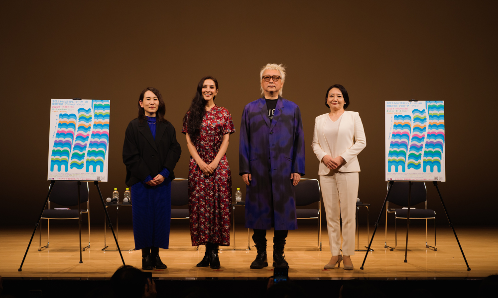 「開館２年前プロローグイベント」で行ったトークイベントのいち場面です。舞台上には、イベント出演者４人が横一列に立っています。出演者は、左から、相馬千秋、サヘル・ローズ、やない道彦、せいけ愛です。その左右には、ｍチルダｍを波模様のようにデザインしたポップな印象のポスターがイーゼルで立てられています。その後ろには、黒いパイプ椅子が数脚のほかに、ペットボトルが数個置かれたサイドテーブルがあります。舞台上は暗めの照明で照らされています。