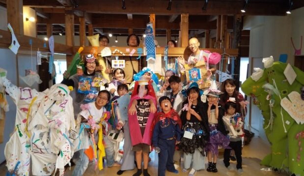 みなみのしえさんが過去に行ったワークショップが終わったあとの集合写真です。子どもたちを中心に１９人が室内に集まっています。全員が工作で作った衣装や小道具を身につけています。紙や布で作られたカラフルなマントや飾り、大きな緑の着ぐるみなどが見え、会場全体がにぎやかな雰囲気です。